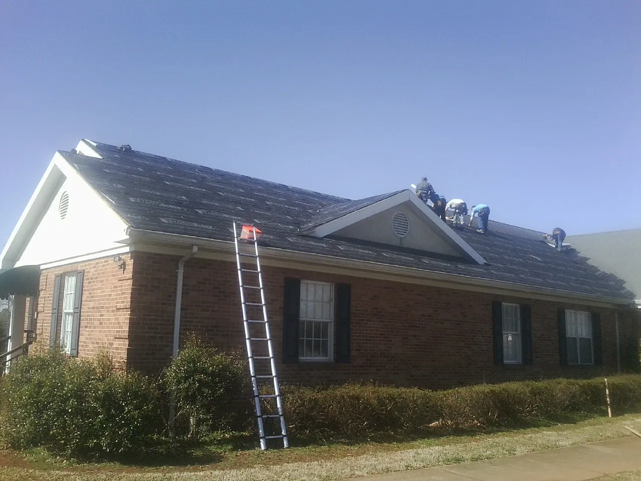 Crew tearing shingles off a brick ranch roof with a ladder set — Metal Roof Repair work by our crew in Lake Park