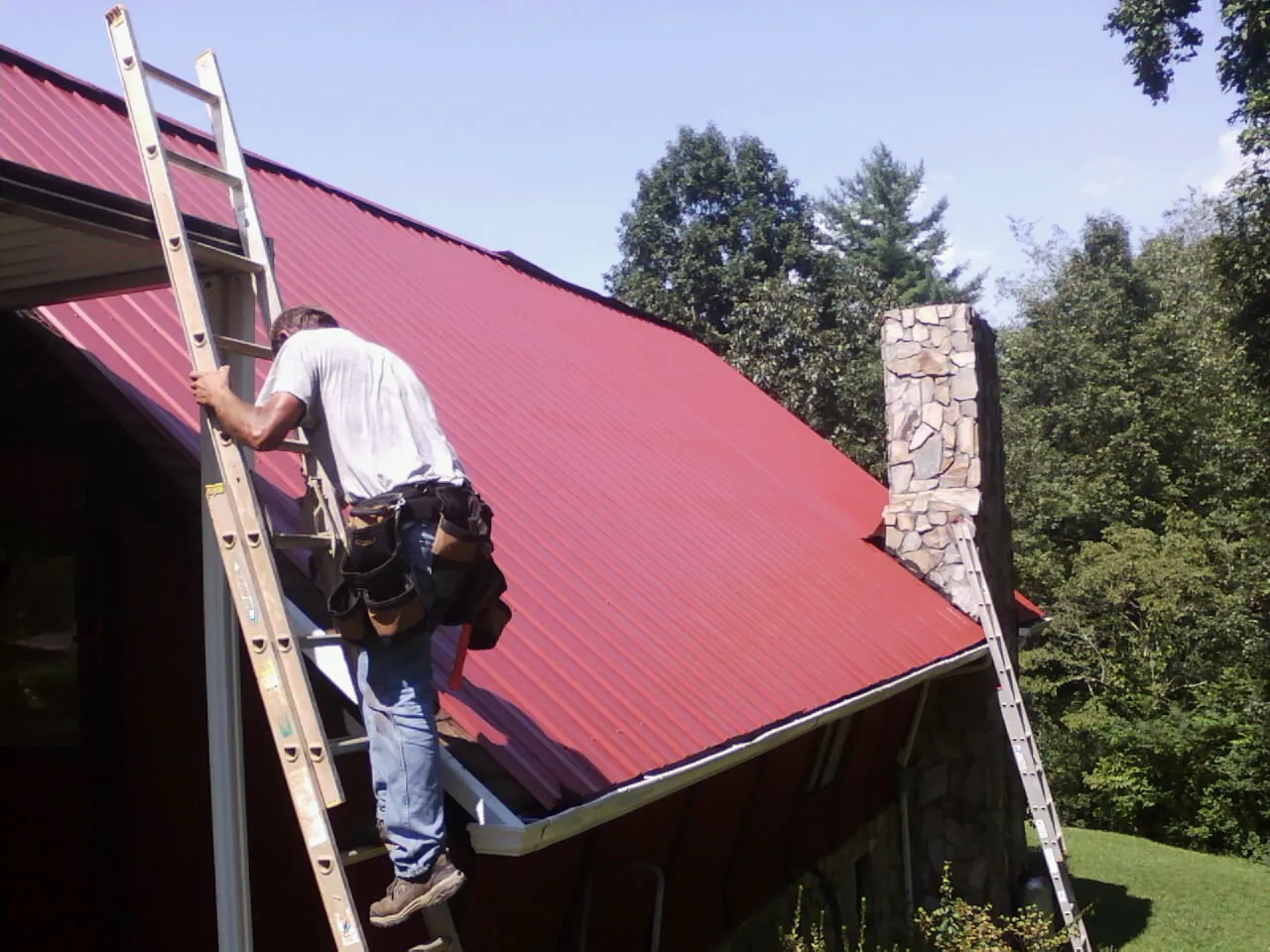 Roofer on a ladder beside a red metal roof and stone chimney — Soffit & Fascia Repair by Roofing Brothers in Lake Park